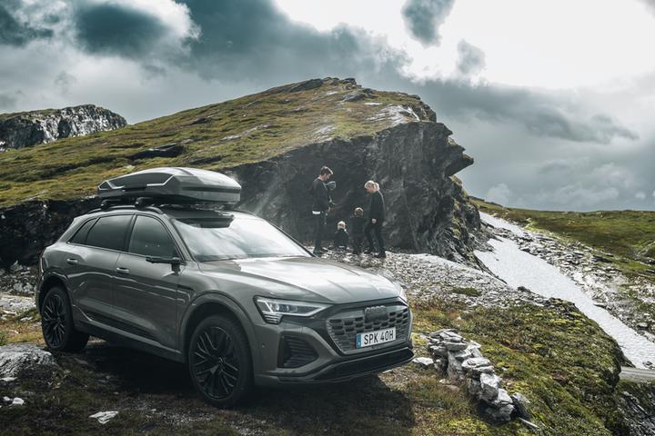 Audi SUV parked on a rocky mountain trail with Thule Motion 3 L roof cargo box and family standing nearby under cloudy sky