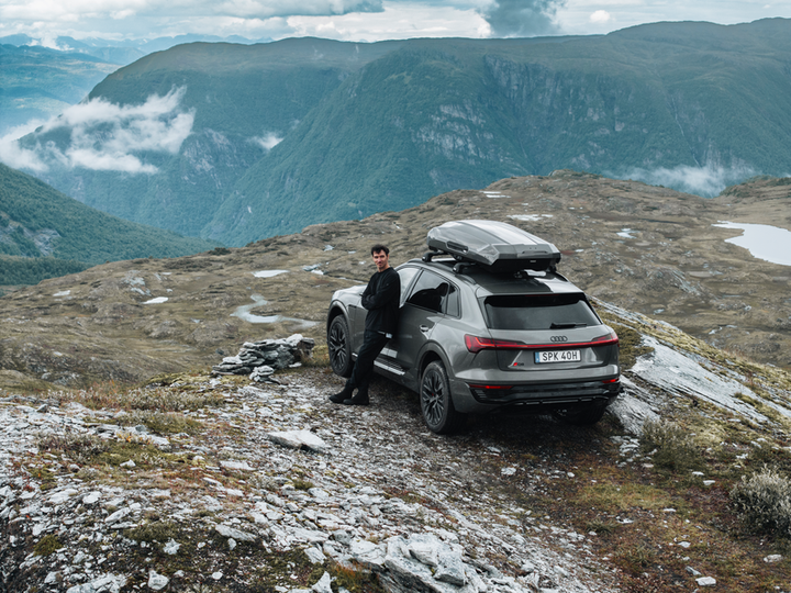 Thule Motion 3 XL rooftop cargo box mounted on a gray SUV with a person standing beside it in a mountainous landscape.